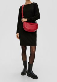 Sac à épaule en cuir rouge avec une forme arrondie, fermeture éclair diagonale et une bandoulière à motifs. Porté avec une robe noire et des bottines noires.