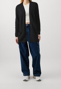 Svart lång cardigan med öppen front, ribbade ärmslut och två framfickor, i kombination med högmidjade blå denimbyxor och vita sneakers.