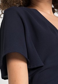 Námořnické modré šaty s texturovanou tkaninou, krátkými, volnými rukávy a výstřihem do V, které zdůrazňují jemný sklonek a hladký povrch.