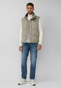 Gepolsterte graue Weste mit Knöpfen, hohem Kragen und Logo-Patch, getragen über einem weißen Rollkragenpullover, kombiniert mit blauen Jeans und beigen Sneakers.