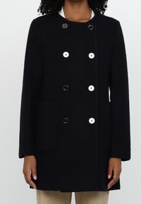 Femme portant un manteau noir à double boutonnage avec des boutons alternés noirs et blancs et deux poches avant, portant un pantalon beige visible en dessous.