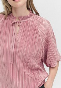 Vaaleanpunainen pleated-paita, jossa on seisova kaulus ja nauhat yksityiskohtana, lyhyet hihat ja teksturoitu pinta. Kankaassa on vaakasuoria raitoja.