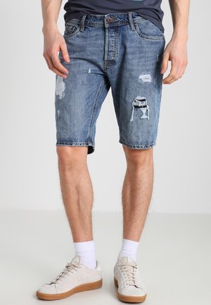 Mand iført midt-lange blå denimshorts med slidte pletter, hvide sokker og lyse beige sneakers, stående på en ensfarvet baggrund.