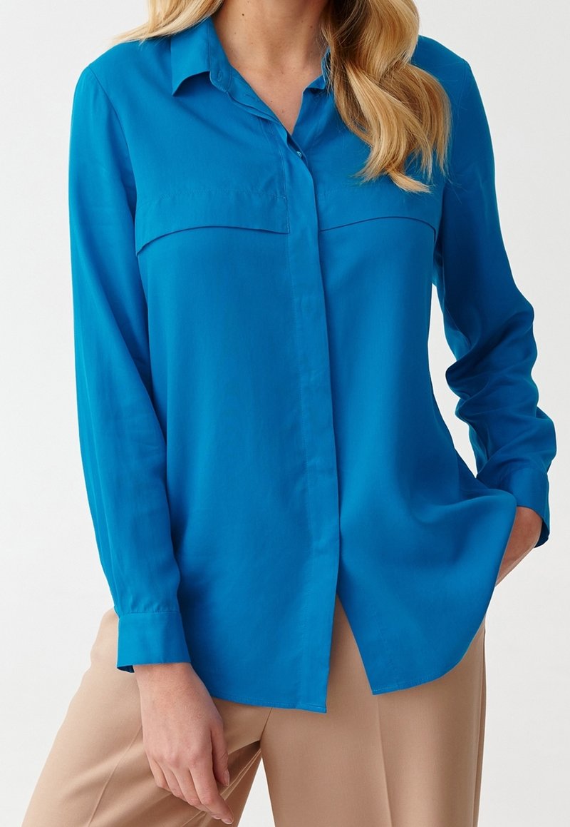 Blå langærmet skjorte med krave, forlommer og knaplukning. Glat tekstur, afslappet pasform og let buet kant.