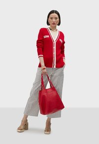 Red cardigan with white stripes, button closure. Paired with striped wide-leg pants, gold-heeled sandals, and a textured red tote bag.