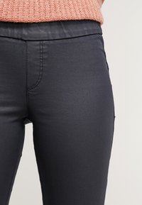 Pantaloni neri elasticizzati con una texture liscia e un design aderente, caratterizzati da tasche laterali e una vita a media altezza.
