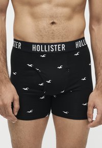 Мъжки торс, облечен в черни боксерки Hollister с бели лого на чайки и марка на колана, стоящ с ръце до тялото.