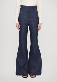 Mörk denim med utsvängda byxor med hög midja, med kontrasterande orange sömmar och sidofickor. Slät textur och breda ben.