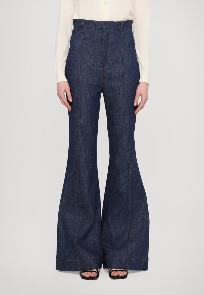 Mörk denim med utsvängda byxor med hög midja, med kontrasterande orange sömmar och sidofickor. Slät textur och breda ben.