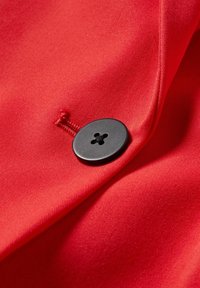 Bouton noir à quatre trous cousu sur un tissu rouge avec des points de couture visibles et une texture pliée autour du bouton.