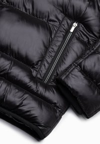 Chaqueta tipo puff negra con textura acolchada, que presenta un bolsillo con cremallera lateral y un tejido interior en contraste. El material es brillante y suave.