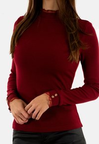 Maglietta a maniche lunghe ribbed color bordeaux con collo alto, caratterizzata da accenti a volant e dettagli in oro sui polsini.