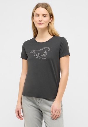 Frau mit langen blonden Haaren, die ein schwarzes T-Shirt mit einer weißen Grafik eines laufenden Pferdes und dem Text "MUSTANG" trägt, kombiniert mit hellgrauen Hosen.