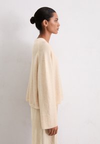 Pull en tricot beige avec des manches larges et un ourlet côtelé, associé à une jupe côtelée assortie, vue de profil contre un fond uni.
