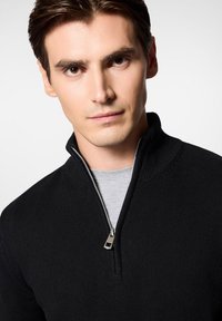 Czarny sweter z zamkiem błyskawicznym o prążkowanej fakturze i wysokim kołnierzu. Posiada srebrny zamek i jest noszony na szarym T-shircie.