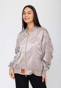 Vrouw glimlachend, draagt een lichtgrijze satijnen bomberjas met ritssluiting aan de voorkant, geborduurde "B" op de borst, ritsvak op de mouw en een oranje label dicht bij de zoom.