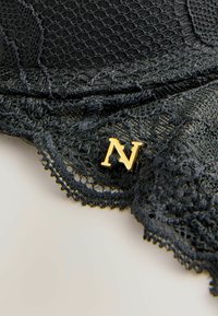 Il tessuto in pizzo nero presenta motivi intricati con bordi scallop. Un accento dorato a forma di lettera "N" è attaccato, aggiungendo dettaglio al design.