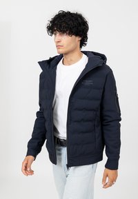 Marineblauer gepolsterter Mantel mit Reißverschluss, abnehmbarem Kapuze und Seitentaschen. Mit einem Logo auf der Brust und strukturierten Stoffakzenten.