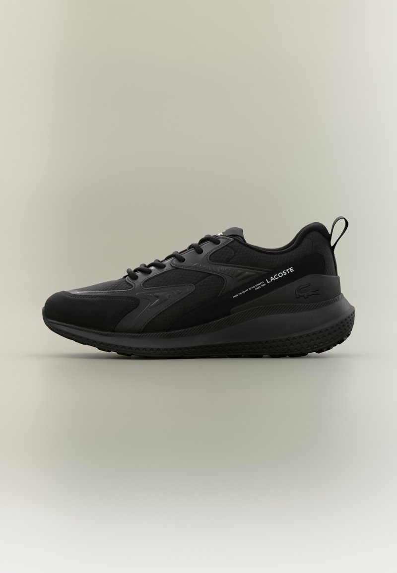 Baskets de sport noires Lacoste avec design à lacets, semelle texturée et logo sur le côté et sur la languette du talon, présentées sur un fond neutre.
