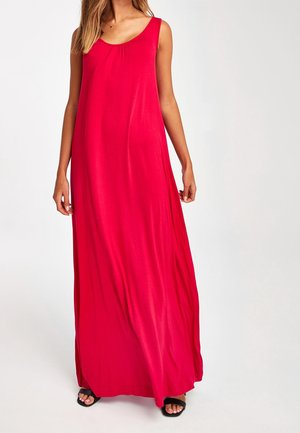 Femme portant une robe rouge sans manches, ample et longue jusqu'au sol, avec un décolleté en U et des sandales noires à bout ouvert.