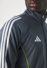 Mørkegrå atletisk jakke med høj krave, hvide striber på skuldrene og limegrønne syersømmer. Har en lynlåslukning foran.