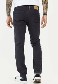 Donkerblauwe denim jeans met vijf zakken, een leren merklabel op de tailleband en een taps toelopende pasvorm. Draagt met witte en marineblauwe sneakers.