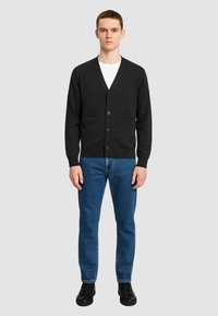 Cardigan nero con scollo a v, bottoni frontali e due tasche applicate, abbinato a jeans blu e scarpe nere su uno sfondo semplice.
