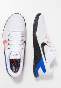 Zapatillas de entrenamiento Nike blancas con parte superior texturizada, suela negra y acentos en azul y naranja intensos. Presentan un cierre de cordones y una plantilla con patrón.