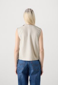 Ärmellose beige Bluse mit Kragen, aus leichtem Stoff gefertigt, kombiniert mit blauen Jeans aus Denim mit einer Stichdetail an der Gesäßtasche.