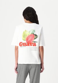 Valge puuvillane t-särk, millel on guava graafika rohelises ja punases värvitoonis, seljal on sõna "Guava" julge punase tekstiga. Ribaline alläär.
