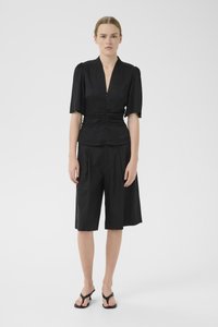 Black short-sleeve blouse with a V-neck and gathered waist, paired with knee-length black pleated shorts and black flip-flops.