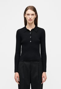 Γυναίκα με ίσια καστανά μαλλιά, που φοράει στενό ribbed μαύρο πόλο πουκάμισο με μακριά μανίκια και τέσσερα κουμπιά, καθώς και μαύρο παντελόνι με ψηλή μέση.