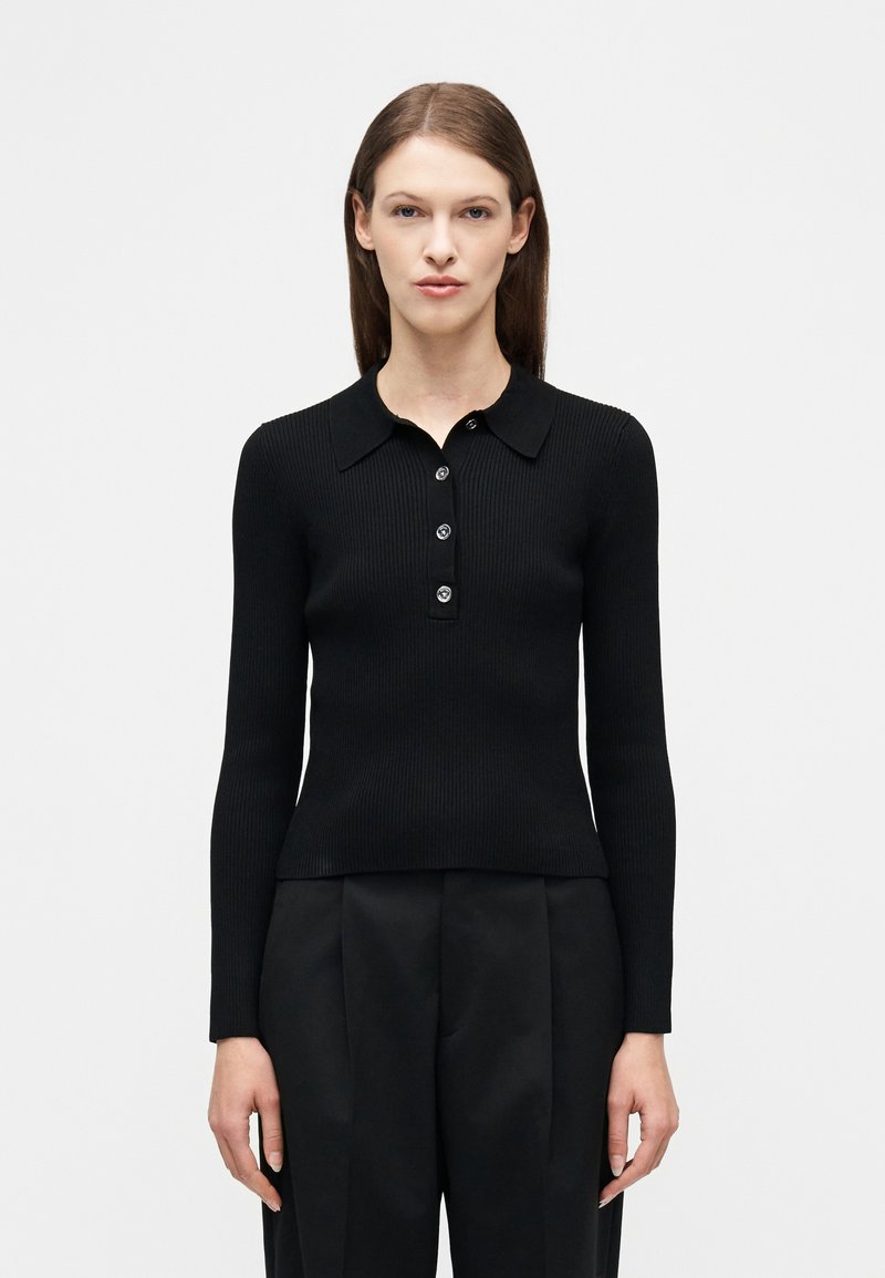 Γυναίκα με ίσια καστανά μαλλιά, που φοράει στενό ribbed μαύρο πόλο πουκάμισο με μακριά μανίκια και τέσσερα κουμπιά, καθώς και μαύρο παντελόνι με ψηλή μέση.