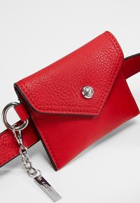 Petite pochette en cuir rouge avec rabat et bouton argenté sur une ceinture assortie, chaîne argentée et étiquette attachées à la boucle de la ceinture.