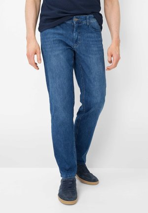 Mann trägt blaue gerade geschnittene Jeans, marineblaue Sneaker und ein dunkelblaues Hemd vor einem schlichten weißen Hintergrund.