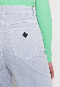 Calças de sarja azul claro com uma superfície texturizada, apresentando uma etiqueta de marca preta em forma de diamante no bolso traseiro. Parte superior verde de mangas longas visível.