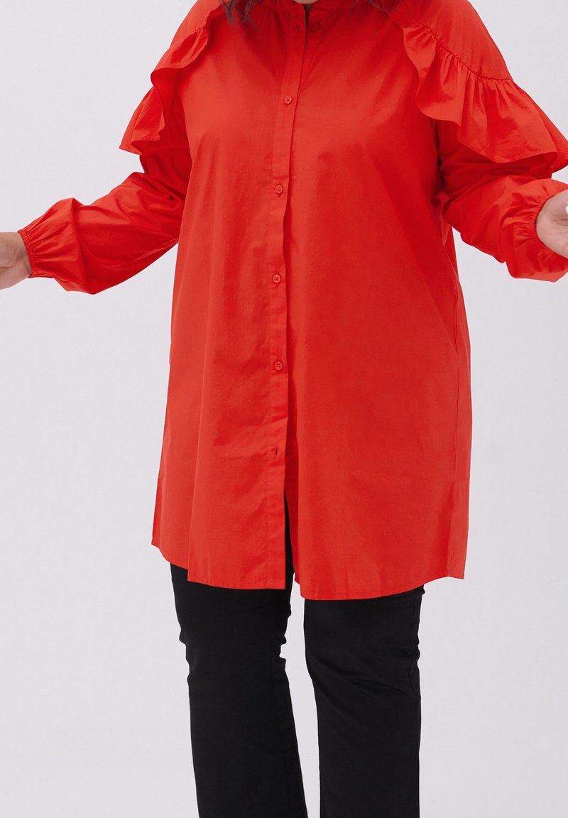 Camicia rossa con bottoni e maniche arricciate, realizzata in tessuto liscio, abbinata a pantaloni neri. La camicia presenta un taglio dritto e spacchi laterali.