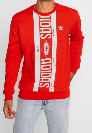 Osoba nosící červenou mikinu Adidas s bílými svislými pruhy a výrazným logem, doplněnou světle modrými džínami.