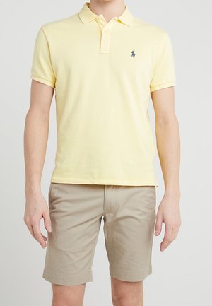 Mann trägt ein hellgelbes kurzärmeliges Poloshirt und hellbeige knielange Shorts und steht vor einem einfarbigen Hintergrund.