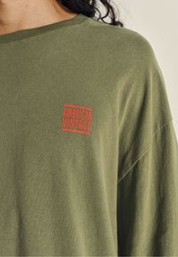 Oliven grønn oversized t-shirt lavet af blødt stof, med et rødt "AMERICAN VINTAGE" logo patch på brystet. Rund halsudskæring og afslappet pasform.
