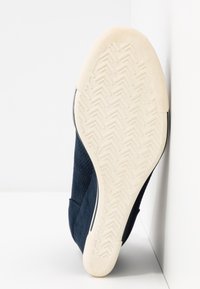 Semelle en caoutchouc blanche avec motif à chevrons sur une basket bleu foncé reposant contre un coin de mur et de sol blancs.