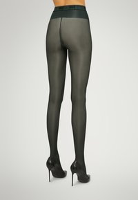 Collant opachi verde scuro con una texture liscia, caratterizzati da un'ampia fascia in vita e abbinati a tacchi alti neri lucidi.