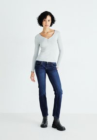 Gray long-sleeve shirt with a V-neck and button detail paired with dark blue jeans and black ankle boots, against a light background.