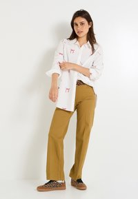 Chemise blanche à boutons avec un imprimé de homards rouges ; pantalons larges de couleur moutarde ; chaussures à imprimé léopard avec une semelle marron.