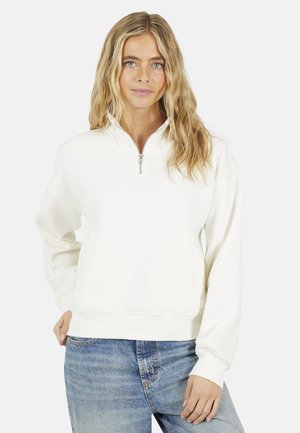 Donna con lunghi capelli biondi che indossa una felpa bianca con zip parziale e jeans blu, in piedi davanti a uno sfondo chiaro e semplice.