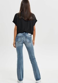 Femme aux longs cheveux bruns portant un tee-shirt noir, un jean en denim bleu et des baskets blanches, se tenant de dos devant un fond uni.