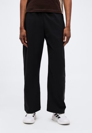 Personne portant un pantalon noir large, des baskets blanches avec des rayures noires, et une chemise marron foncé avec des bandes latérales beiges.