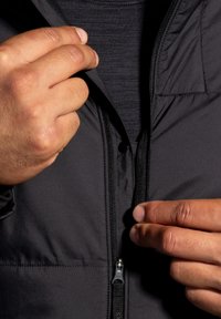 Giacca nera imbottita con chiusura a zip. Presenta una superficie liscia e un colletto alto. Primo piano delle mani che regolano la cerniera.
