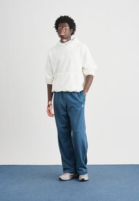Sweat à capuche blanc avec lettrage texturé, associé à un pantalon de sport bleu avec des bandes blanches sur les côtés et des baskets grises.
