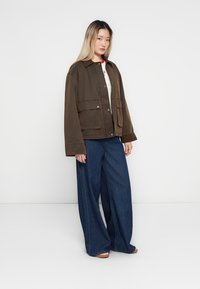 Hnědá oversized bunda s knoflíkovým zapínáním a dvěma předními kapsami, v kombinaci s volnými modrými džíny, na modelce proti jednobarevnému pozadí.